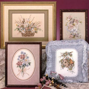 May include: A collection of framed floral cross-stitch art pieces. The pieces include a basket of flowers, a bouquet, and a birdhouse scene. The frames are various colors, including gold, brown, and a light blue pillow with a floral design.