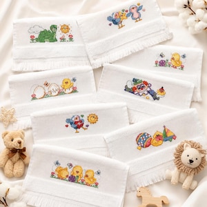 Puede incluir: Una colección de toallas de mano rectangulares blancas con bordes de flecos, cada una con un diseño único de punto de cruz colorido que incluye un dinosaurio verde, pájaros, un payaso, pollitos y juguetes, junto a un pequeño oso y león de peluche.