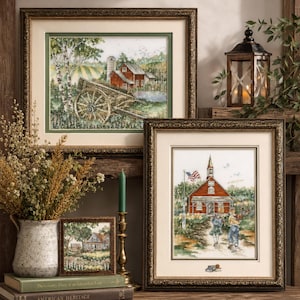 May include: A collection of framed landscape art pieces with gold ornate frames. The art depicts rural scenes with red buildings, trees, and figures. A lantern and vase with dried flowers are also present on a wooden shelf.