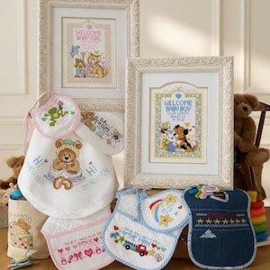 May include: A collection of baby-themed items, including framed cross-stitch art with "Welcome Baby Girl" and "Welcome Baby Boy" messages. Bibs with various designs, such as a teddy bear, frog, and a truck, are also present. The color palette includes white, pink, blue, and yellow.