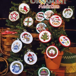 Puede incluir: Una colección de adornos navideños bordados, cada uno con un diseño único, incluyendo un cardenal rojo, un muñeco de nieve y un pingüino. Los adornos se exhiben en un pequeño árbol de Navidad, con el texto "ORNAMENT Addiction" en la parte superior.
