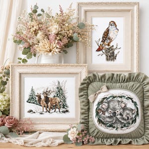 Könnte beinhalten: Präsentation von gerahmten Kreuzsticharbeiten und einem dekorativen Kissen. Die gerahmten Stücke zeigen eine Winterlandschaft mit Hirschen, einen Falken und Koalas. Eine Vase mit Trockenblumen ergänzt die Szene.