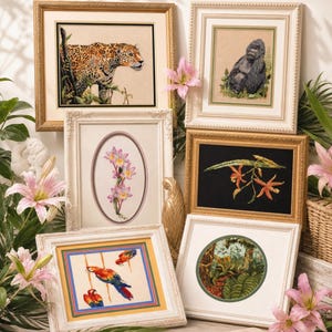 May include: A collection of framed needlepoint art featuring various wildlife and botanical themes. The artwork includes a leopard, gorilla, parrots, orchids, and water lilies. Frames are gold and white, with a variety of shapes and sizes.