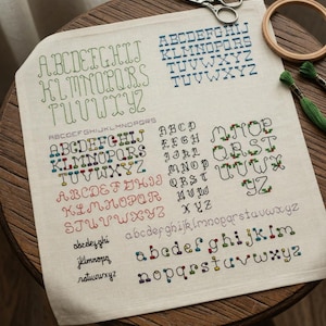 May include: A linen fabric displays various embroidered alphabets in different fonts and colors. The sampler includes uppercase and lowercase letters in green, blue, red, and black threads. A wooden embroidery hoop and scissors are visible.