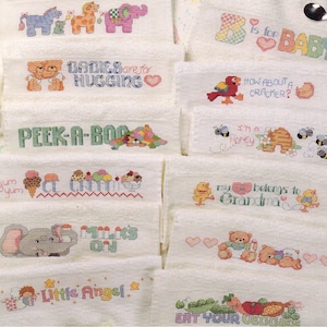 May include: A collection of white baby washcloths with cross-stitched designs. Each cloth features a different image and text, including animals, ice cream, and phrases like "BABIES are for HUGGING" and "EAT YOUR VEGGIES".