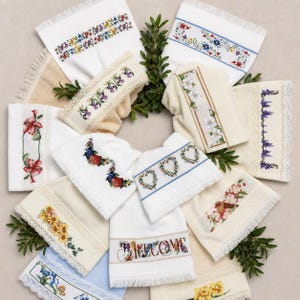 May include: A collection of white and cream-colored hand towels featuring various floral cross-stitch patterns, lace trim, and fringe edges. One towel displays the word WELCOME in a floral font, while others show birds, hearts, and blossoms.