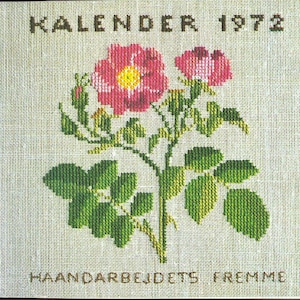 Puede incluir: Página de calendario de punto de cruz de 1972 con un diseño floral. La imagen muestra dos rosas rosas con centros amarillos, hojas verdes y el texto "KALENDER 1972" y "HAANDARBEJDETS FREMME". El diseño está sobre una tela beige claro.