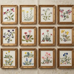 May include: Twelve framed floral cross-stitch artworks. Each piece showcases a different flower design in various colours, set against a white background. The frames are ornate, gold-coloured, and rectangular, creating a vintage aesthetic.