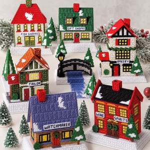 May include: A collection of handmade, colorful, miniature houses, each with a unique design and shop name, including "Fish Shoppe," "Gift Shoppe," and "Bakery." The houses are decorated with snow and Christmas trees, creating a festive village scene.