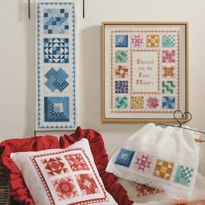 May include: A collection of cross-stitch items featuring quilt block designs. Includes a framed piece with the text "Blessed are the Piece Makers", a pillow, a hanging banner, and a drawstring bag, all in various colors.