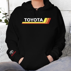 May include: Black hooded sweatshirt with the word "TOYOTA" in white block letters above a horizontal stripe in yellow, orange, and red. The Toyota logo and text are on the left sleeve. The sweatshirt has a front pocket.