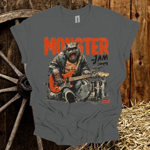 May include: A gray t-shirt with a distressed graphic featuring a monster playing a red electric guitar and drums. The text "MONSTER JAM" is printed in orange above the illustration. The shirt has a classic crew neck.