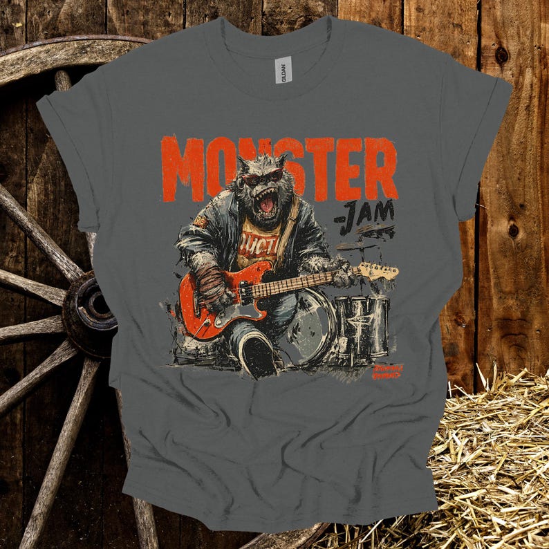 May include: A gray t-shirt with a distressed graphic featuring a monster playing a red electric guitar and drums. The text "MONSTER JAM" is printed in orange above the illustration. The shirt has a classic crew neck.