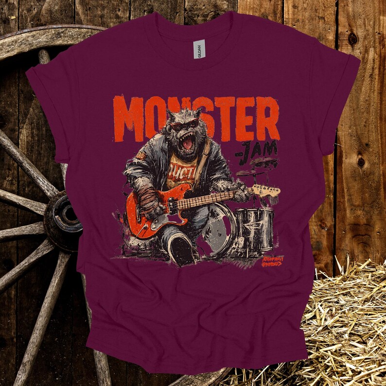 May include: Burgundy t-shirt with a graphic of a monster playing a red guitar and drums. The text "MONSTER JAM" is above the monster. The shirt has a crew neck and short sleeves. The graphic is in shades of orange, gray, and black.