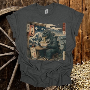 May include: Dark gray t-shirt featuring a Godzilla graphic enjoying ramen at a Japanese restaurant. The design includes Japanese text and a scenic background with a mountain and ocean waves. The shirt is made of a soft material.