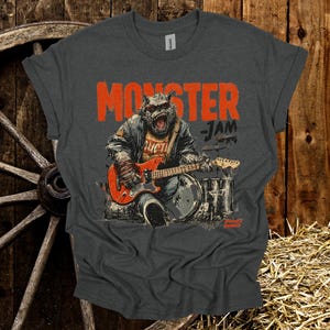 May include: Dark gray t-shirt featuring a monster playing an electric guitar and drums. The shirt has the words "MONSTER JAM" in orange block letters. The monster is wearing a jacket and has a fierce expression.