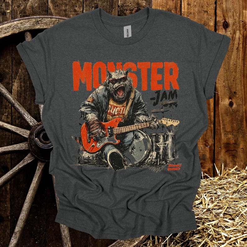 May include: Dark gray t-shirt featuring a monster playing an electric guitar and drums. The shirt has the words "MONSTER JAM" in orange block letters. The monster is wearing a jacket and has a fierce expression.