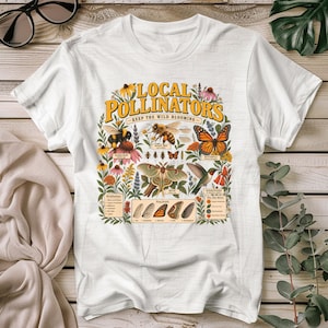 Op de afbeelding: Een wit t-shirt met korte mouwen en een kleurrijke grafische print met de tekst LOCAL POLLINATORS KEEP THE WILD BLOOMING. Het ontwerp bevat gedetailleerde illustraties van een hommel, honingbij, vlinder, mot en kolibrie met wilde bloemen.