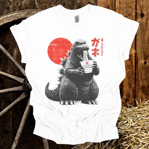 Puede incluir: Camiseta blanca con un gr&aacute;fico de Godzilla comiendo fideos ramen. El dise&ntilde;o incluye un sol rojo y texto japon&eacute;s. La ilustraci&oacute;n de Godzilla es en blanco y negro, con el cuenco de ramen en blanco y rojo.