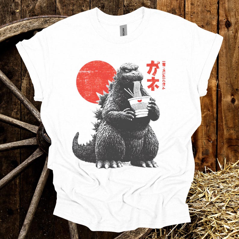 Puede incluir: Camiseta blanca con un gr&aacute;fico de Godzilla comiendo fideos ramen. El dise&ntilde;o incluye un sol rojo y texto japon&eacute;s. La ilustraci&oacute;n de Godzilla es en blanco y negro, con el cuenco de ramen en blanco y rojo.