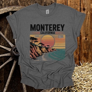 May include: Gray t-shirt featuring a scenic illustration of the California coast with the text "Monterey California." The design includes a sunset over the ocean, cliffs, and trees, creating a vintage travel aesthetic.