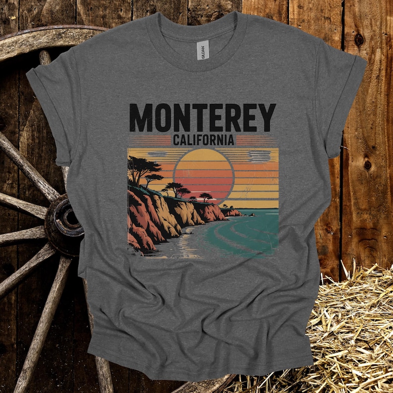May include: Gray t-shirt featuring a scenic illustration of the California coast with the text "Monterey California." The design includes a sunset over the ocean, cliffs, and trees, creating a vintage travel aesthetic.
