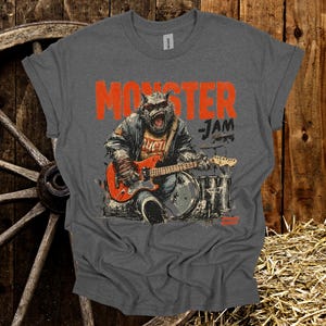May include: Gray t-shirt with the words "MONSTER JAM" in orange block letters. The shirt features a graphic of a monster playing a red electric guitar and drums. The monster is wearing a jacket and has a fierce expression.