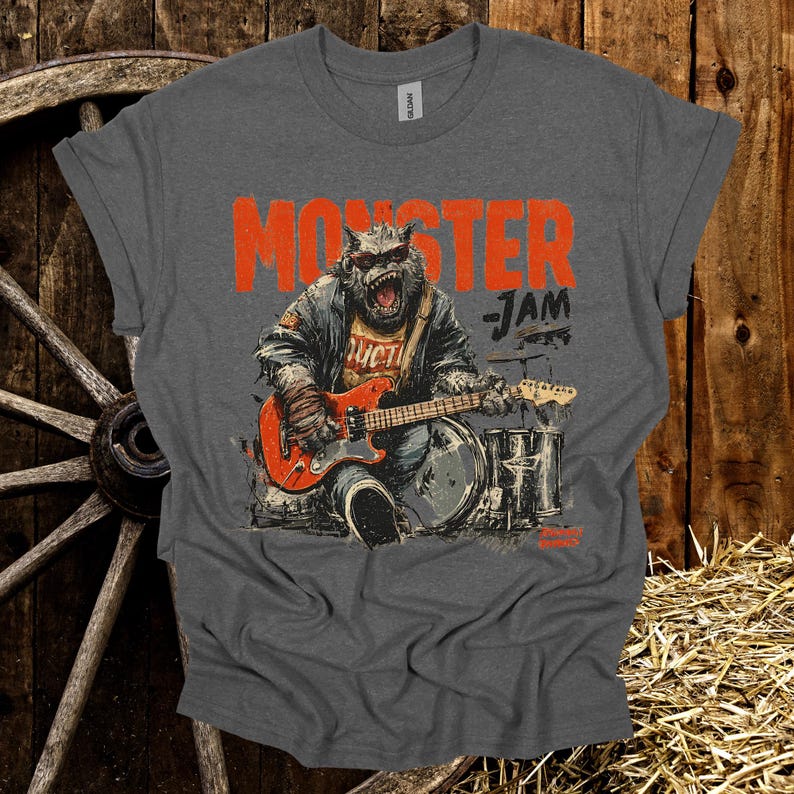 May include: Gray t-shirt with the words "MONSTER JAM" in orange block letters. The shirt features a graphic of a monster playing a red electric guitar and drums. The monster is wearing a jacket and has a fierce expression.