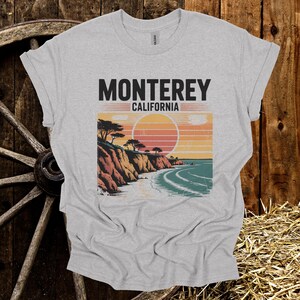 May include: A heather gray t-shirt featuring a coastal landscape design with the text "Monterey California". The graphic depicts a sunset over the ocean with cliffs and trees. The shirt is made of a soft material.