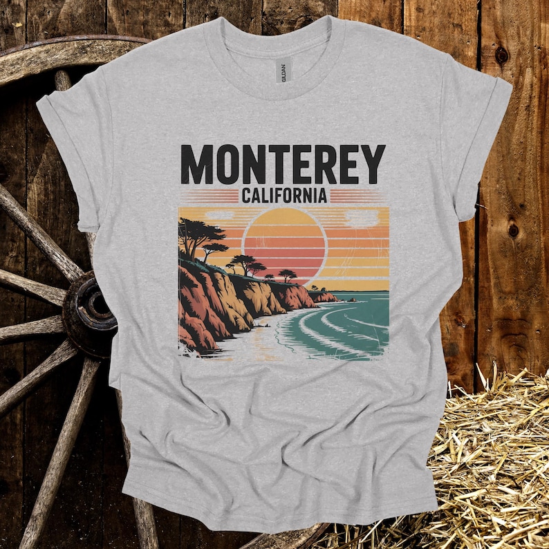 May include: A heather gray t-shirt featuring a coastal landscape design with the text "Monterey California". The graphic depicts a sunset over the ocean with cliffs and trees. The shirt is made of a soft material.
