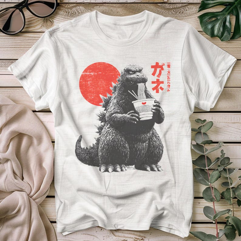 Puede incluir: Camiseta blanca con un dise&ntilde;o de Godzilla comiendo fideos ramen. El dise&ntilde;o incluye un sol rojo y texto japon&eacute;s. El gr&aacute;fico es en blanco y negro, con un estilo vintage.