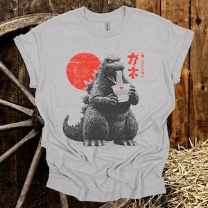 Puede incluir: Camiseta gris claro con un dise&ntilde;o de Godzilla comiendo fideos ramen. Un sol rojo est&aacute; detr&aacute;s de Godzilla, y texto japon&eacute;s est&aacute; a la derecha. La camiseta est&aacute; hecha de un material suave. El dise&ntilde;o est&aacute; impreso al frente.