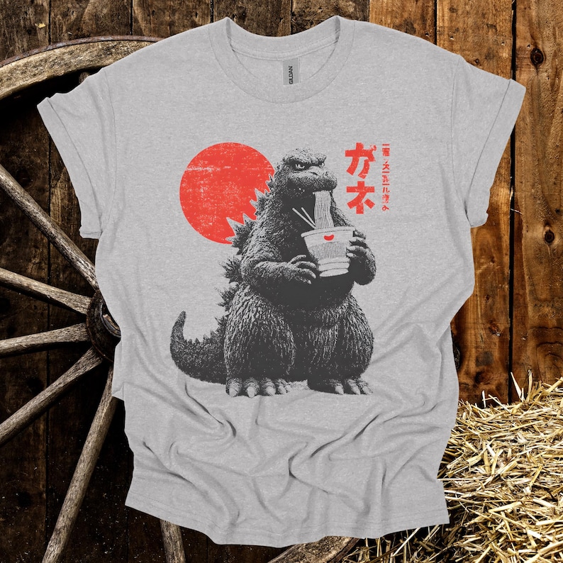 Puede incluir: Camiseta gris claro con un dise&ntilde;o de Godzilla comiendo fideos ramen. Un sol rojo est&aacute; detr&aacute;s de Godzilla, y texto japon&eacute;s est&aacute; a la derecha. La camiseta est&aacute; hecha de un material suave. El dise&ntilde;o est&aacute; impreso al frente.