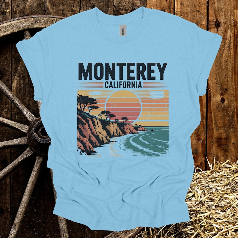 May include: Light blue t-shirt featuring a graphic of the California coast with the text "Monterey California". The design includes a sunset, cliffs, trees, and ocean waves. The shirt is made of a soft material.