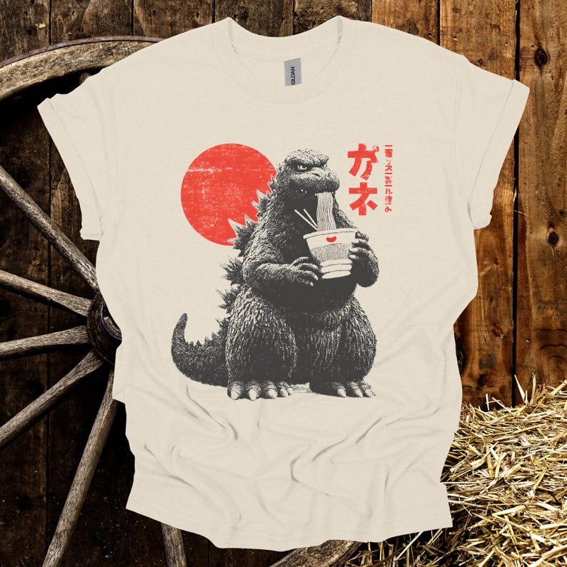 Puede incluir: Camiseta color crema con un gr&aacute;fico de Godzilla comiendo fideos ramen. El dise&ntilde;o incluye un sol rojo y texto japon&eacute;s. El gr&aacute;fico est&aacute; en tonos grises y negros.