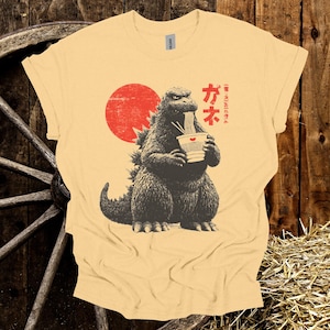 Puede incluir: Camiseta amarillo claro con un gr&aacute;fico de Godzilla comiendo fideos ramen. El dise&ntilde;o incluye un sol rojo y texto japon&eacute;s. La camiseta es de la marca Gildan.