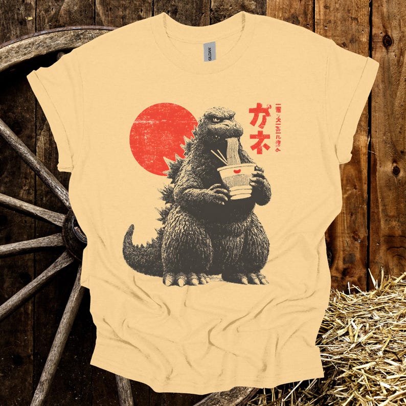Puede incluir: Camiseta amarillo claro con un gr&aacute;fico de Godzilla comiendo fideos ramen. El dise&ntilde;o incluye un sol rojo y texto japon&eacute;s. La camiseta es de la marca Gildan.