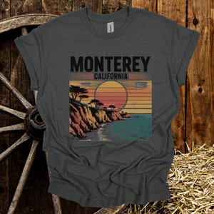 May include: Dark gray t-shirt featuring a graphic of a coastal landscape with the text "Monterey California." The design includes a sunset, cliffs, trees, and ocean. The shirt is made of a soft material.