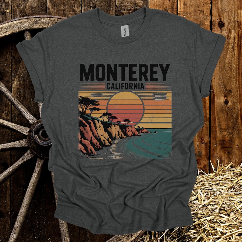 May include: Dark gray t-shirt featuring a graphic of a coastal landscape with the text "Monterey California." The design includes a sunset, cliffs, trees, and ocean. The shirt is made of a soft material.