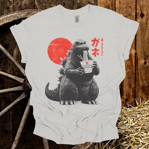 Puede incluir: Camiseta gris claro con un gr&aacute;fico de Godzilla comiendo fideos ramen. El dise&ntilde;o incluye un sol rojo y texto japon&eacute;s. La camiseta es de la marca Gildan.