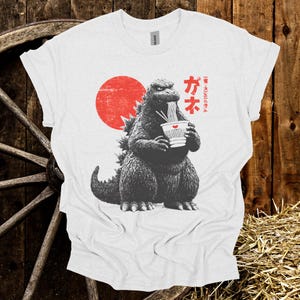 Puede incluir: Camiseta blanca con un dise&ntilde;o de Godzilla comiendo ramen, con un sol rojo de fondo. Texto japon&eacute;s impreso en la camiseta. El dise&ntilde;o utiliza tonos grises y rojos.