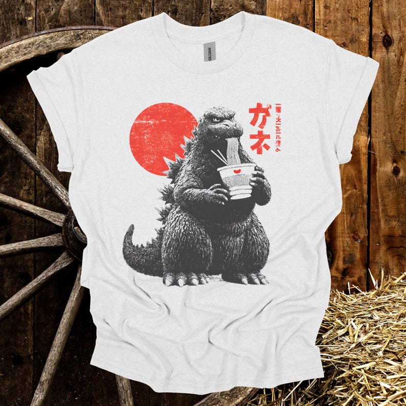 Puede incluir: Camiseta blanca con un dise&ntilde;o de Godzilla comiendo ramen, con un sol rojo de fondo. Texto japon&eacute;s impreso en la camiseta. El dise&ntilde;o utiliza tonos grises y rojos.