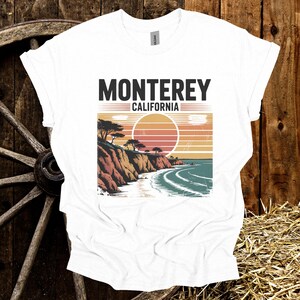 May include: White t-shirt with a graphic of a coastal landscape featuring the text "Monterey California". The design includes a sunset, cliffs, trees, and ocean waves, creating a vintage travel aesthetic.