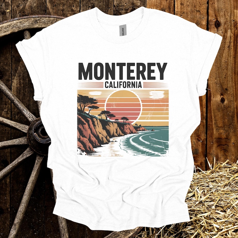 May include: White t-shirt with a graphic of a coastal landscape featuring the text "Monterey California". The design includes a sunset, cliffs, trees, and ocean waves, creating a vintage travel aesthetic.