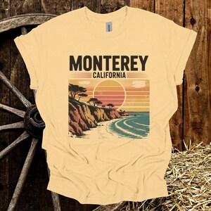 May include: A light yellow t-shirt featuring a graphic of a coastal scene with the text "MONTEREY CALIFORNIA". The design includes a sunset, cliffs, trees, and ocean waves. The shirt is made of a soft material.