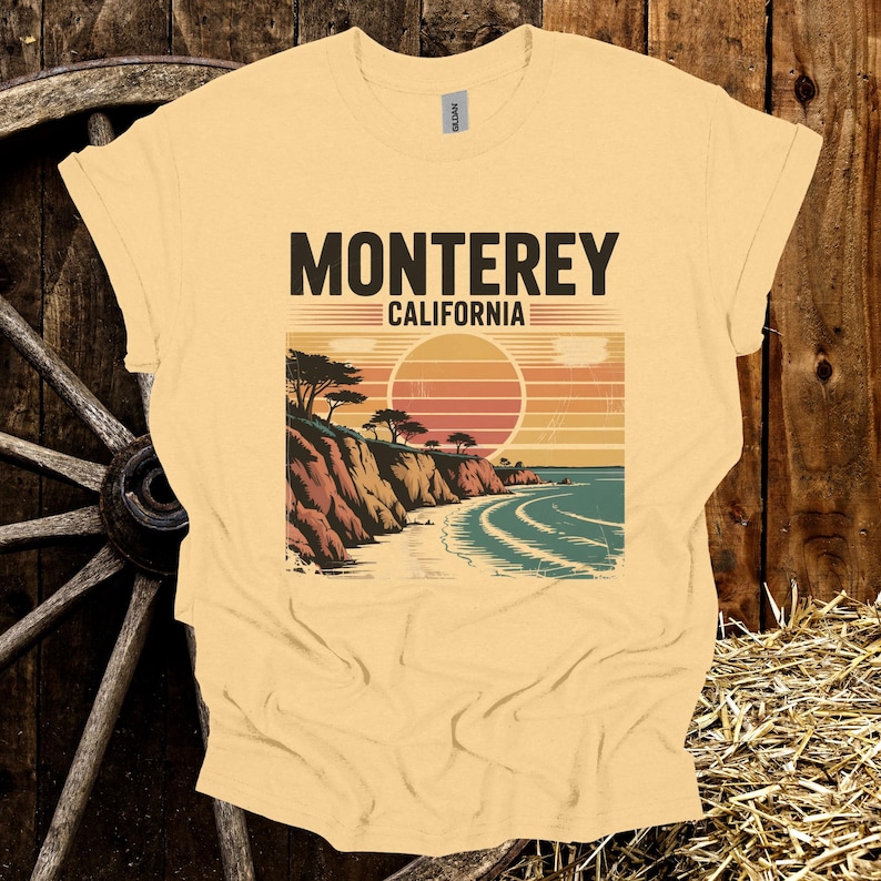 May include: A light yellow t-shirt featuring a graphic of a coastal scene with the text "MONTEREY CALIFORNIA". The design includes a sunset, cliffs, trees, and ocean waves. The shirt is made of a soft material.