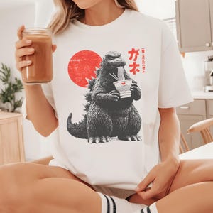 Puede incluir: Camiseta blanca con un dise&ntilde;o de Godzilla en blanco y negro comiendo fideos, con un sol rojo de fondo. Texto japon&eacute;s impreso en la camiseta. La camiseta la lleva una persona.