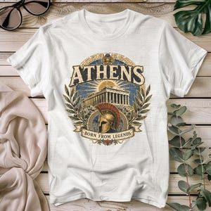 Op de afbeelding: Een wit t-shirt met korte mouwen en een cirkelvormige afbeelding met de tekst ATHENS en BORN FROM LEGENDS. Het ontwerp bevat een gedetailleerde illustratie van het Parthenon, een gouden Spartaanse helm en olijftakken in blauwe en gouden tinten.