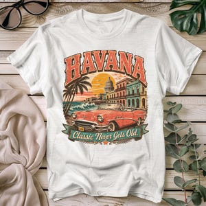 Op de afbeelding: Een wit T-shirt met korte mouwen en een grafische print van een rode vintage cabriolet voor gebouwen in Havana en palmbomen. De tekst HAVANA staat bovenaan en Classic Never Gets Old staat op een banner eronder.