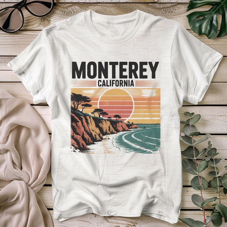 May include: White t-shirt featuring a graphic of a coastal scene with the text "Monterey California." The design includes a sunset, ocean waves, and cliffs with trees. The shirt is on a wooden surface.